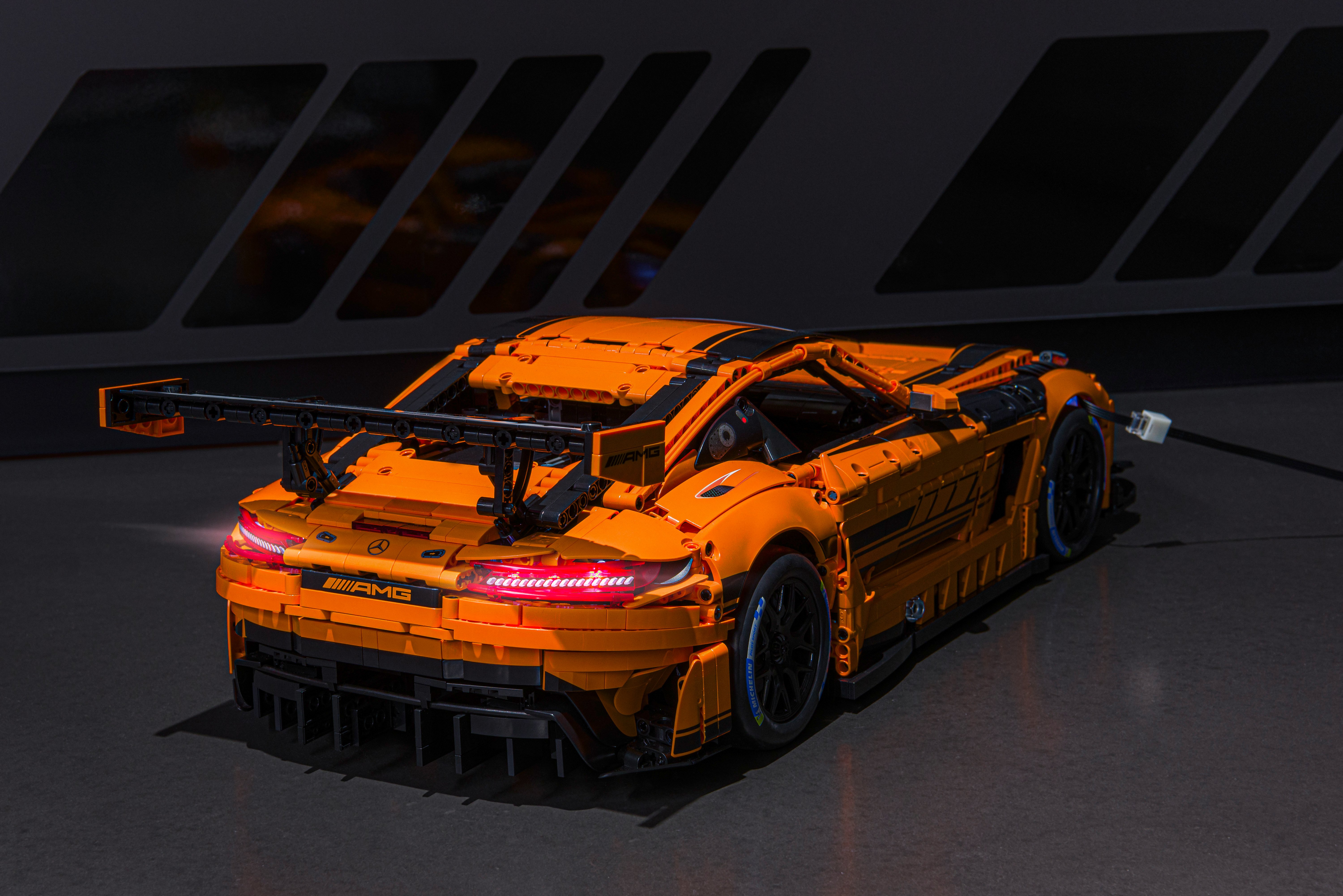 Rückansicht des orangefarbenen Mercedes-AMG GT3 Klemmbaustein-Modells mit eingeschalteten LED-Rückleuchten; sichtbar sind der große Heckflügel, die detaillierte Karosserie sowie das per USB angeschlossene Lichtsystem auf dunklem Untergrund.