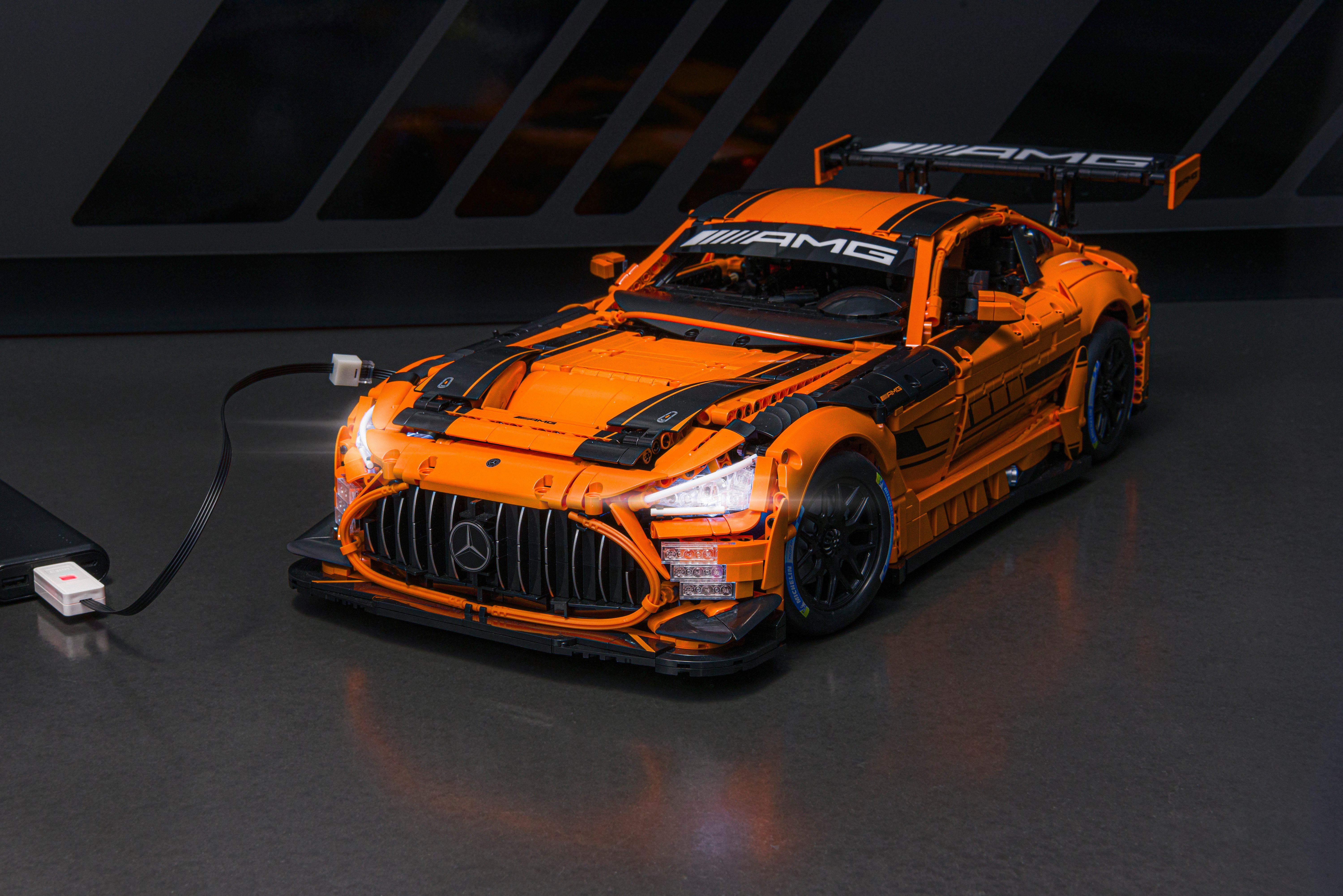 Beleuchtetes Mercedes-AMG GT3 Klemmbaustein-Modell in Orange; die angeschlossenen LED-Scheinwerfer leuchten hell, während das Fahrzeug auf dunklem Untergrund steht und per USB-Kabel mit Strom versorgt wird.
